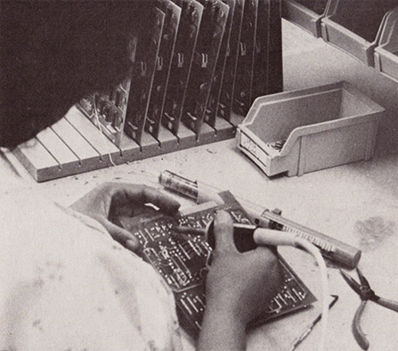 The process of assembling videogames for home or arcade is long and arduous. Here, at Exidy, a technician solders a printed circuit board. Other boards, stacked in a rack to her lefl, await the woman’s attention.